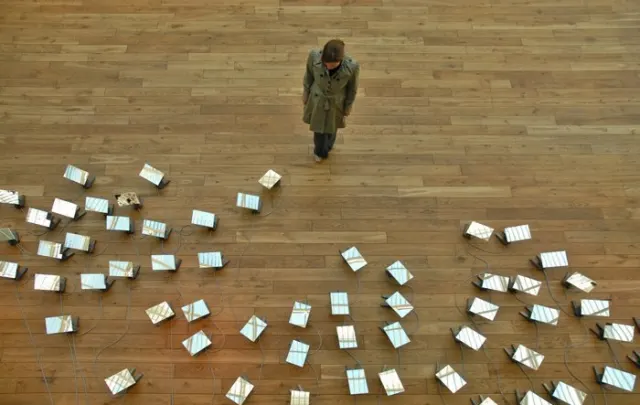Vue de haut d'un parquet clair : une personne en manteau se tient debout au milieu, entour&eacute;e d'une cinquantaine de petits miroirs pos&eacute;s au sol et reli&eacute;s par des c&acirc;bles, tous orient&eacute;s vers elle.
