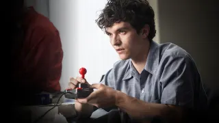 Un jeune homme aux cheveux boucl&eacute;s, assis devant un &eacute;cran hors champ, tient un joystick &agrave; boule rouge des deux mains, le regard fixe.
