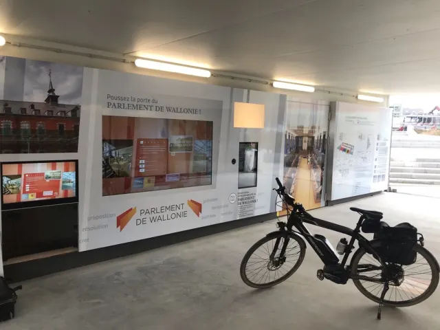 Le couloir en enfilade : la longue paroi imprim&eacute;e avec ses &eacute;crans &agrave; gauche, le sol de b&eacute;ton lisse, un v&eacute;lo &eacute;lectrique noir gar&eacute; au premier plan et une valise pos&eacute;e par terre. Au fond, &agrave; droite, un escalier de b&eacute;ton nu et une structure de chantier.