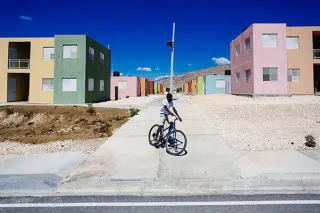 Un enfant &agrave; v&eacute;lo s'arr&ecirc;te au bord d'une rue neuve bord&eacute;e de petits immeubles peints en vert, rose et ocre, sous un ciel bleu