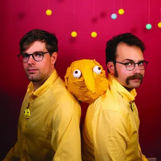 Photo promotionnelle du duo : deux hommes en chemise jaune, dos &agrave; dos sur fond rose, encadrent une marionnette jaune aux gros yeux ronds ; des boules de couleur pendent au-dessus d'eux