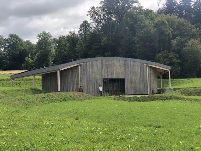 Vu de l'ext&eacute;rieur, un long b&acirc;timent bas au bardage de bois gris&eacute; et au toit de zinc l&eacute;g&egrave;rement incurv&eacute;, &agrave; demi enterr&eacute; dans un talus d'herbe. Une baie vitr&eacute;e sombre marque l'entr&eacute;e, o&ugrave; se tiennent deux personnes. Autour, une prairie fauch&eacute;e en pente et un rideau d'arbres sous un ciel gris.