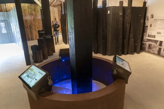 Un bassin circulaire aux parois de m&eacute;tal rouill&eacute;, rempli d'une lumi&egrave;re bleue mouvante, autour d'une large poutre de bois carbonis&eacute; qui monte du centre vers le plafond. Deux pupitres inclin&eacute;s portant chacun un &eacute;cran et un combin&eacute; d'&eacute;coute sont pos&eacute;s sur le rebord. Au fond, la for&ecirc;t de poteaux noircis et une paroi imprim&eacute;e d'une photographie de ruines ; un visiteur s'avance dans l'all&eacute;e.