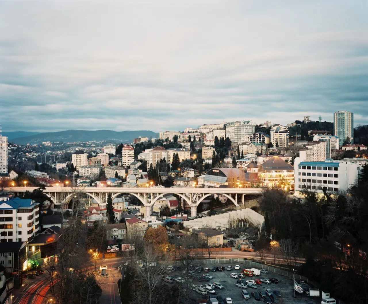 Vue de Sotchi au cr&eacute;puscule : un viaduc &agrave; arches blanches traverse un vallon, des immeubles blancs et des toits de t&ocirc;le escaladent la colline jusqu'aux montagnes, un parking &eacute;clair&eacute; au premier plan.