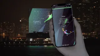 Une main tient un t&eacute;l&eacute;phone o&ugrave; nage un poisson dessin&eacute; ; derri&egrave;re, de l'autre c&ocirc;t&eacute; du port de Hong Kong, la fa&ccedil;ade LED du mus&eacute;e M+ affiche le m&ecirc;me &eacute;tang la nuit