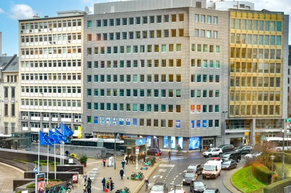 Fa&ccedil;ade d'un immeuble de bureaux des ann&eacute;es soixante au rond-point Schuman, &agrave; Bruxelles. Au premier &eacute;tage, des panneaux bleus &eacute;pellent &laquo; EXPERIENCE EUROPE &raquo; en grandes lettres blanches, et d'autres portent &laquo; VISIT US! &raquo;. Au pied de l'immeuble, un bus, une bouche de m&eacute;tro, des v&eacute;los partag&eacute;s, des passants et une rang&eacute;e de drapeaux europ&eacute;ens sur des m&acirc;ts.