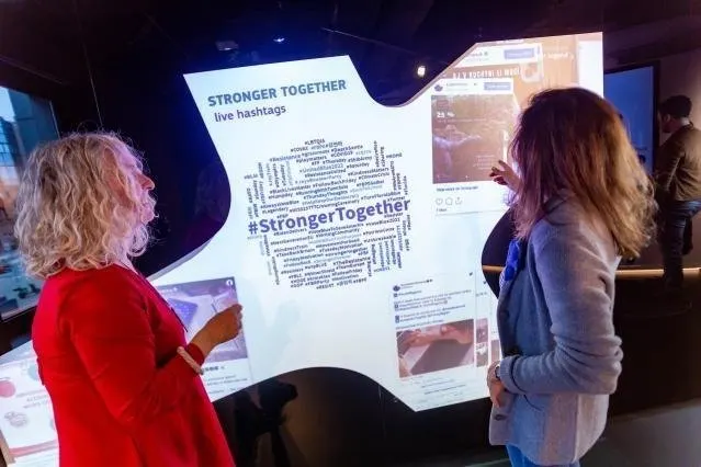 Deux femmes debout de dos et de profil devant une grande surface lumineuse aux bords anguleux. Sur celle-ci, le titre &laquo; STRONGER TOGETHER &mdash; live hashtags &raquo; et un nuage de mots-di&egrave;se de tailles vari&eacute;es autour du mot-di&egrave;se central &laquo; #StrongerTogether &raquo;. &Agrave; droite du nuage, des captures de publications de r&eacute;seaux sociaux avec photos et compteurs de mentions.