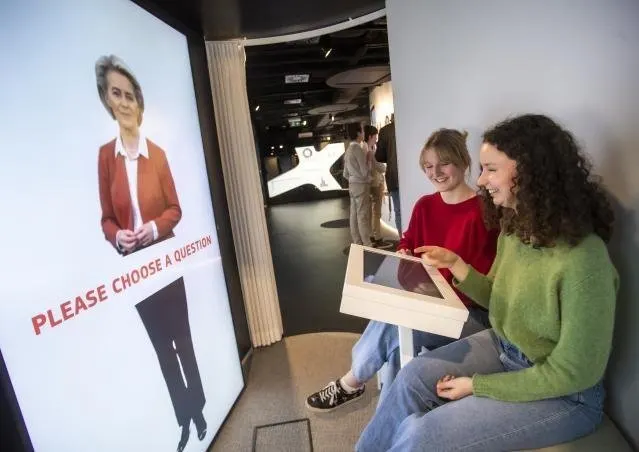 Deux adolescentes assises sur une banquette basse, souriantes, une tablette blanche inclin&eacute;e pos&eacute;e sur les genoux de l'une d'elles. Face &agrave; elles, un grand &eacute;cran vertical montre en pied une femme en veste rouge, les mains jointes ; sur l'&eacute;cran, en lettres rouges inclin&eacute;es, &laquo; PLEASE CHOOSE A QUESTION &raquo;. Au fond, un couloir sombre avec d'autres &eacute;crans.