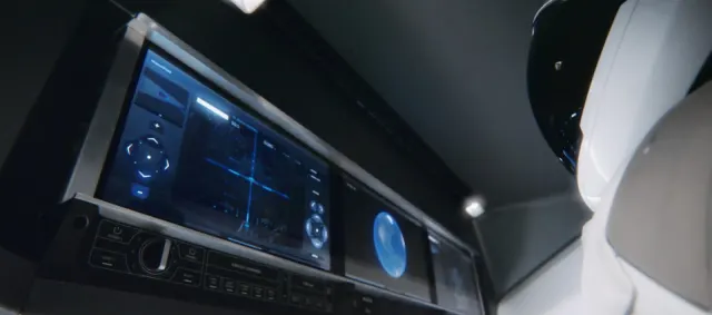 Vue en contre-plong&eacute;e de l'int&eacute;rieur sombre d'une capsule spatiale. Un bandeau de trois &eacute;crans est suspendu au-dessus des si&egrave;ges. Le premier affiche une interface bleu fonc&eacute; : une croix directionnelle &agrave; gauche, une vue centrale avec un r&eacute;ticule et un sch&eacute;ma d'amarrage, des cadrans ronds &agrave; droite. Le deuxi&egrave;me montre la Terre, un petit globe bleu sur fond noir. Sous les &eacute;crans, une rang&eacute;e de commandes physiques : un bouton rotatif, des touches carr&eacute;es, les mentions &laquo; POWER 1 &raquo;, &laquo; RESET &raquo; et &laquo; EXECUTE COMMAND &raquo;. &Agrave; droite, l'&eacute;paule blanche d'une combinaison.